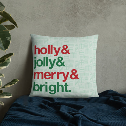 A 22" by 22" throw pillow decorated with a pale green background pattern of presents. There are also four lines of red and green text on the pillow that read "Holly & jolly & merry & bright". The pillow is  set on top of a bed with a blue blanket against a concrete wall.
