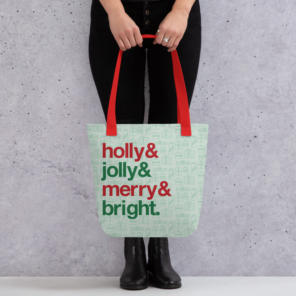 A waist-down shot of a person holding a tote bag with red handles and a printed pattern of light green presents of various sizes. There are also four lines of red and green text on the bag that read "Holly & jolly & merry & bright"