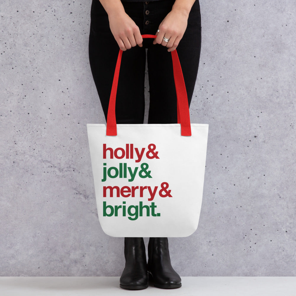 A waist-down shot of a person holding a white tote bag with red handles, decorated with four lines of red and green text. The text reads "Holly & jolly & merry & bright."