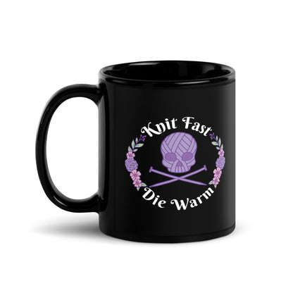 A black 11 ounce mug featuring an image of a purple skull and crossbones made of yarn and knitting needles. A floral wreath surrounds the skull, along with words that read "Knit Fast, Die Warm"
