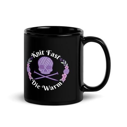 A black 11 ounce mug featuring an image of a purple skull and crossbones made of yarn and knitting needles. A floral wreath surrounds the skull, along with words that read "Knit Fast, Die Warm"