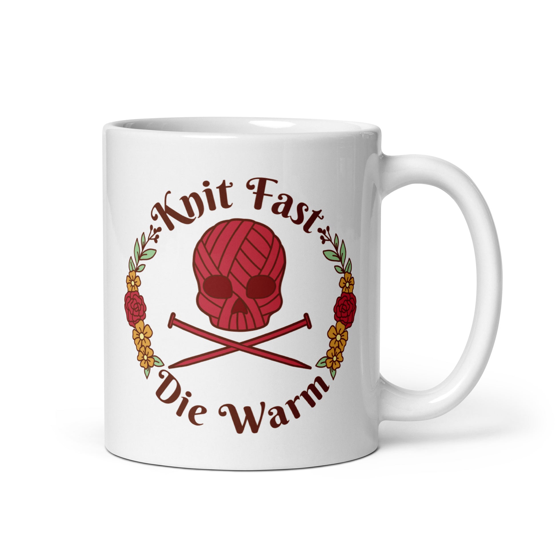 A white 11 ounce mug featuring an image of a red skull and crossbones made of yarn and knitting needles. A floral wreath surrounds the skull, along with words that read "Knit Fast, Die Warm"