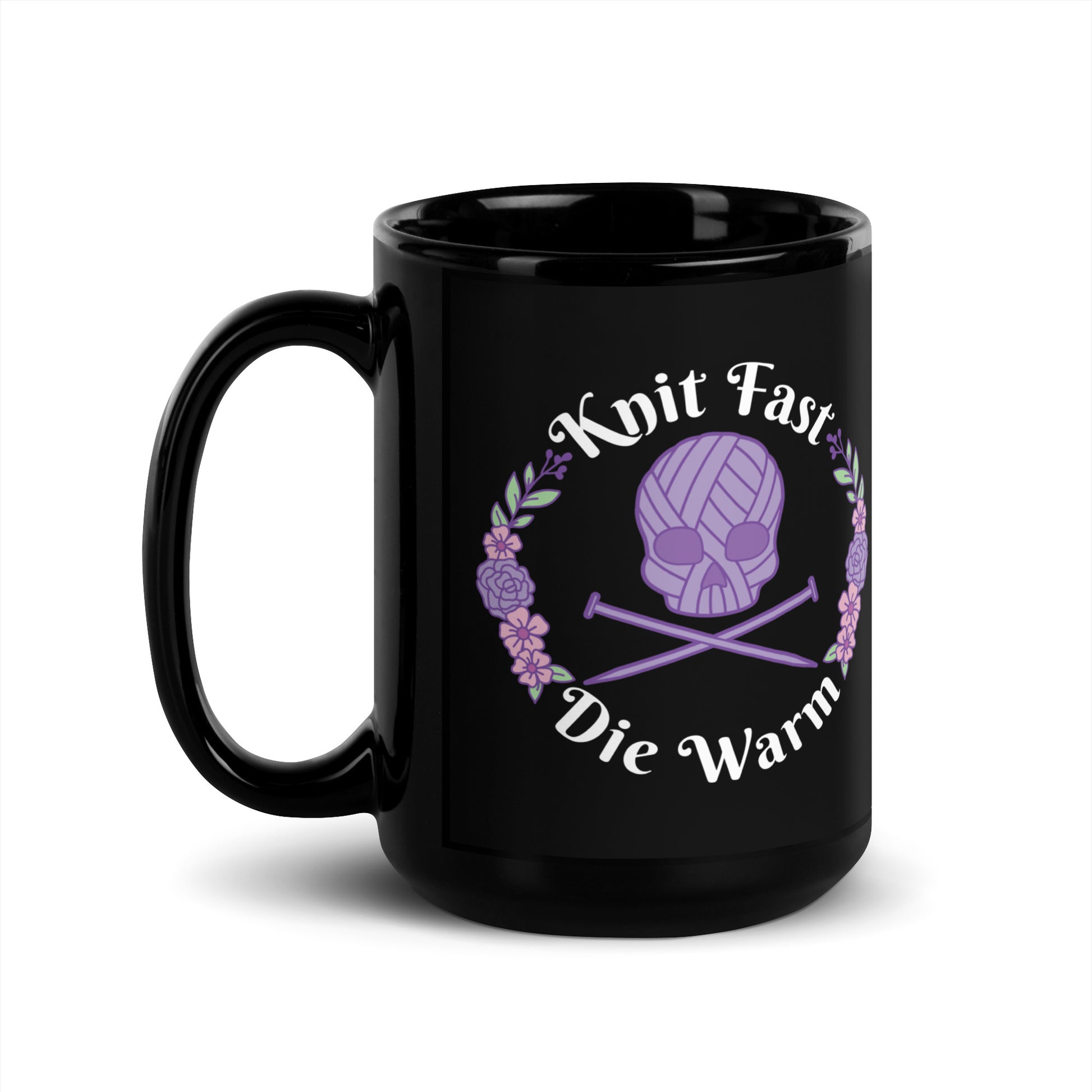 A black 15 ounce mug featuring an image of a purple skull and crossbones made of yarn and knitting needles. A floral wreath surrounds the skull, along with words that read "Knit Fast, Die Warm"
