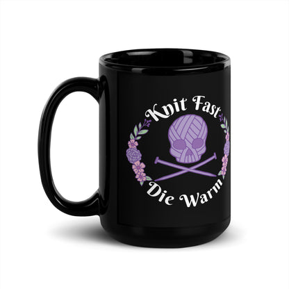 A black 15 ounce mug featuring an image of a purple skull and crossbones made of yarn and knitting needles. A floral wreath surrounds the skull, along with words that read "Knit Fast, Die Warm"