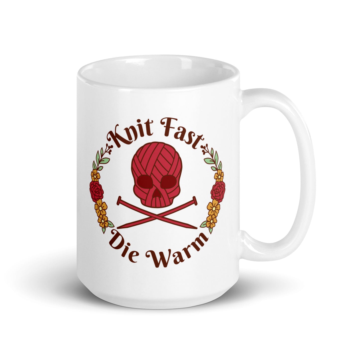 A white 15 ounce mug featuring an image of a red skull and crossbones made of yarn and knitting needles. A floral wreath surrounds the skull, along with words that read "Knit Fast, Die Warm"