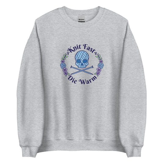 A gray crewneck sweatshirt featuring an image of a skull and crossbones made of yarn and knitting needles. A floral wreath surrounds the skull, along with words that read "Knit Fast, Die Warm"