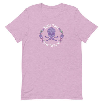 A heathered pink crewneck t-shirt featuring an image of a skull and crossbones made of yarn and knitting needles. A floral wreath surrounds the skull, along with words that read "Knit Fast, Die Warm"