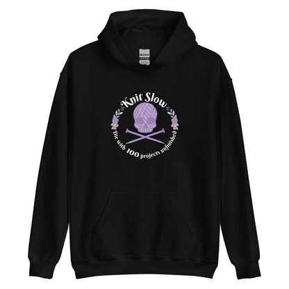 A black hooded sweatshirt featuring an image of a skull and crossbones made of yarn and knitting needles. A floral wreath surrounds the skull, along with words that read "Knit Slow, Die with 100 projects unfinished"