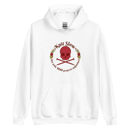 A white hooded sweatshirt featuring an image of a skull and crossbones made of yarn and knitting needles. A floral wreath surrounds the skull, along with words that read "Knit Slow, Die with 100 projects unfinished"