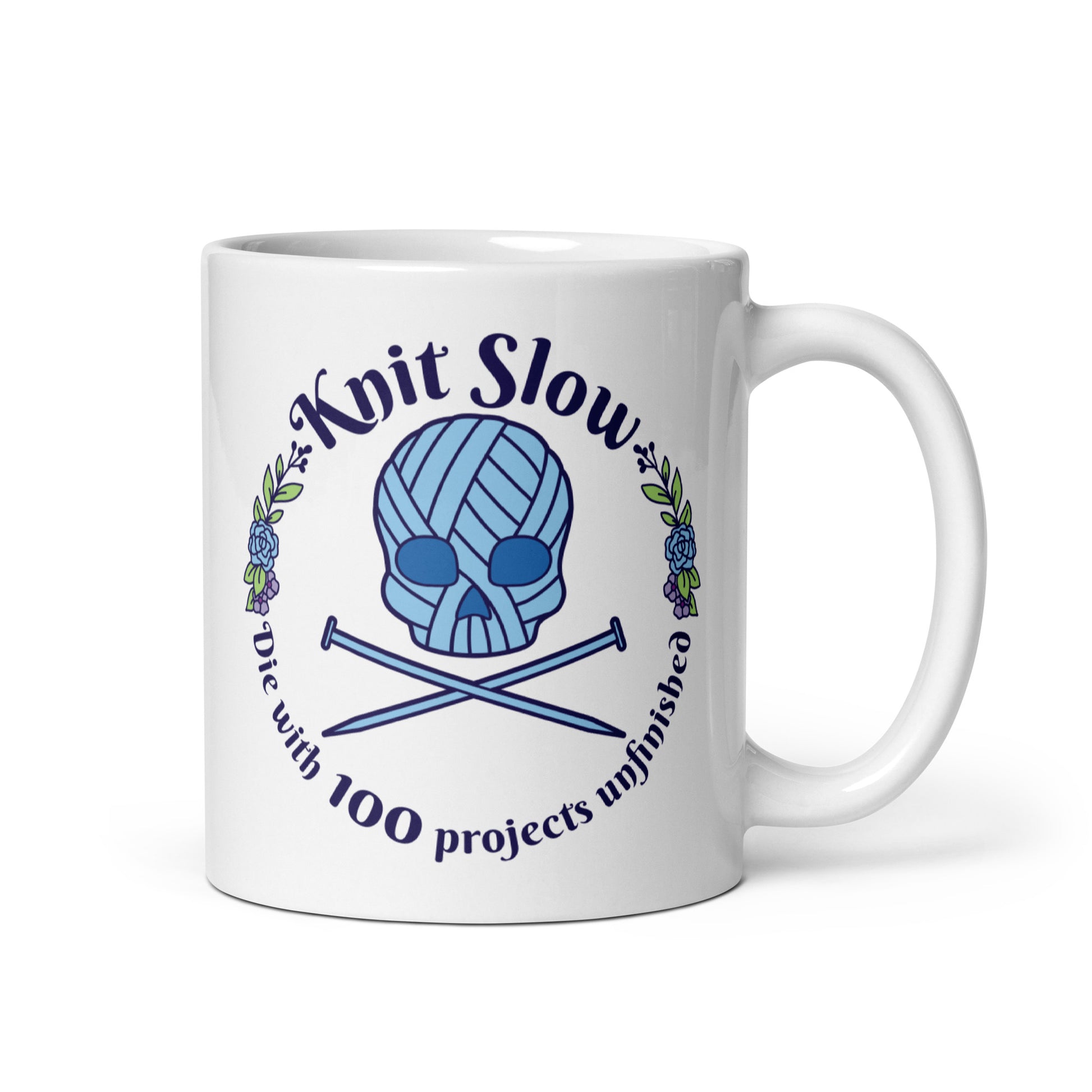 A white 11 ounce ceramic mug featuring an image of a skull and crossbones made of yarn and knitting needles. A floral wreath surrounds the skull, along with words that read "Knit Slow, Die with 100 projects unfinished"
