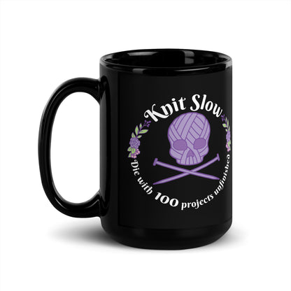 A black 15 ounce ceramic mug featuring an image of a skull and crossbones made of yarn and knitting needles. A floral wreath surrounds the skull, along with words that read "Knit Slow, Die with 100 projects unfinished"
