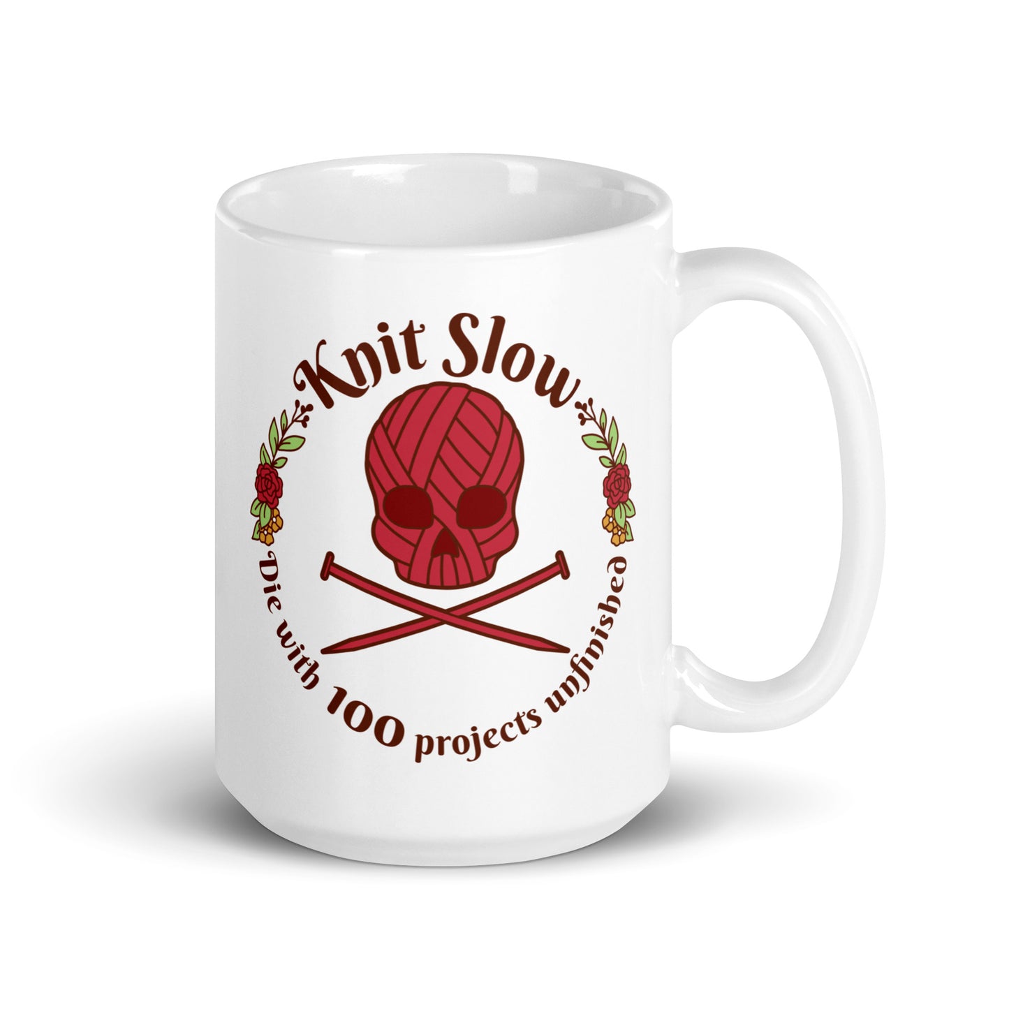 A white 15 ounce ceramic mug featuring an image of a skull and crossbones made of yarn and knitting needles. A floral wreath surrounds the skull, along with words that read "Knit Slow, Die with 100 projects unfinished"
