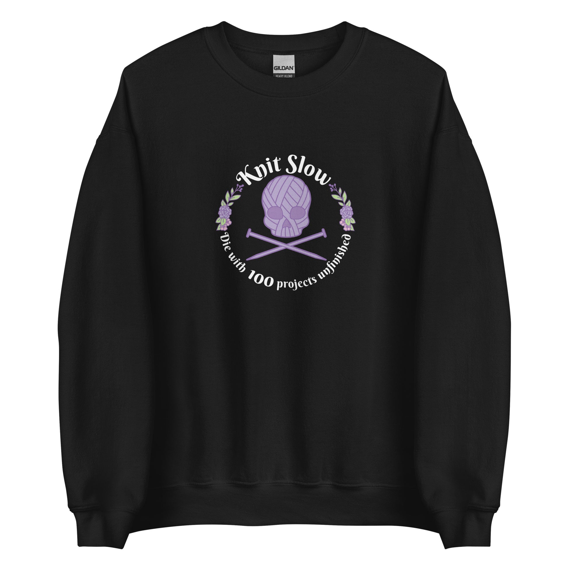 A black crewneck sweatshirt featuring an image of a skull and crossbones made of yarn and knitting needles. A floral wreath surrounds the skull, along with words that read "Knit Slow, Die with 100 projects unfinished"
