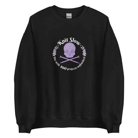 A black crewneck sweatshirt featuring an image of a skull and crossbones made of yarn and knitting needles. A floral wreath surrounds the skull, along with words that read "Knit Slow, Die with 100 projects unfinished"