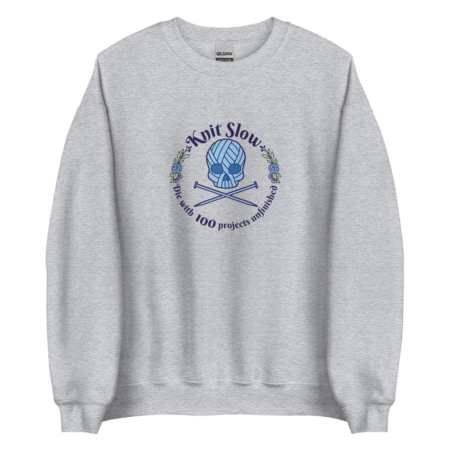 A grey crewneck sweatshirt featuring an image of a skull and crossbones made of yarn and knitting needles. A floral wreath surrounds the skull, along with words that read "Knit Slow, Die with 100 projects unfinished"