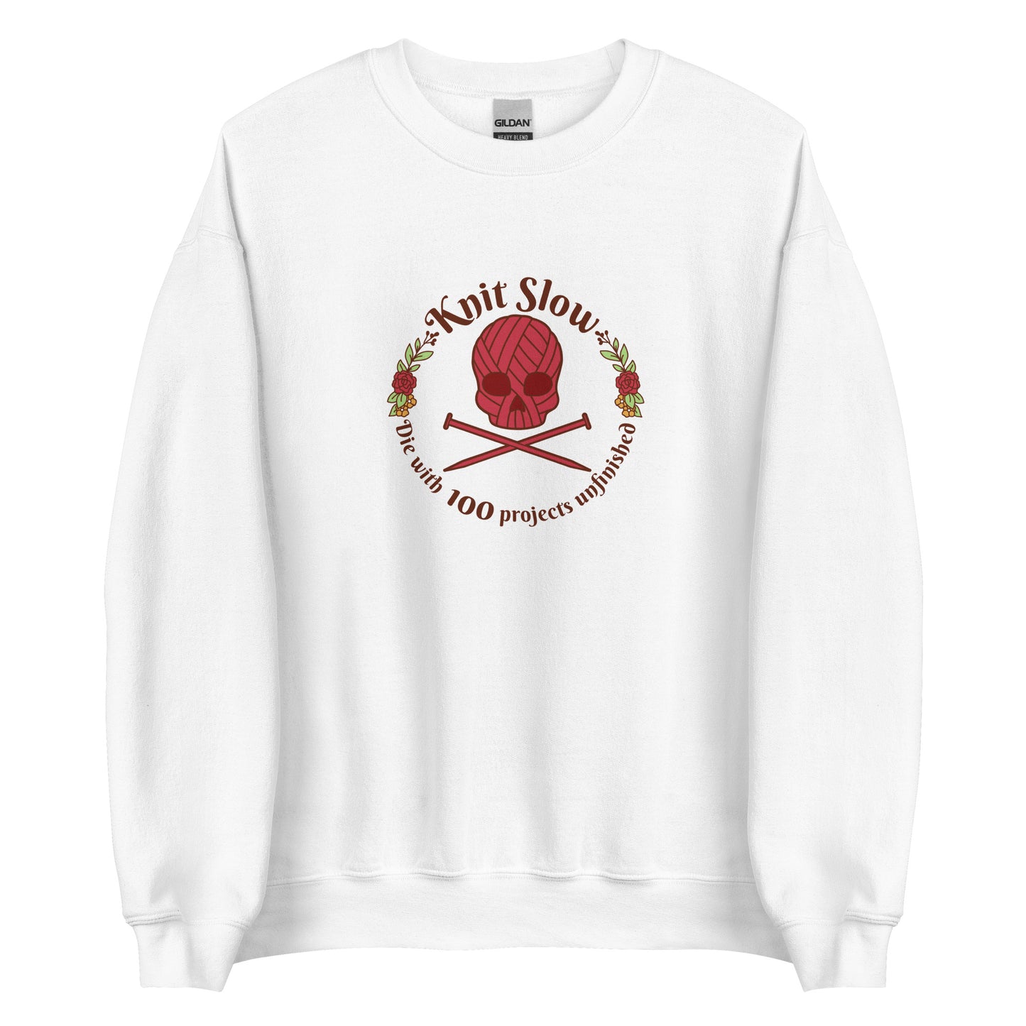 A white crewneck sweatshirt featuring an image of a skull and crossbones made of yarn and knitting needles. A floral wreath surrounds the skull, along with words that read "Knit Slow, Die with 100 projects unfinished"
