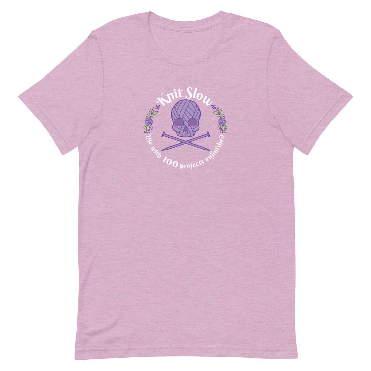 A heathered pink crewneck t-shirt featuring an image of a skull and crossbones made of yarn and knitting needles. A floral wreath surrounds the skull, along with words that read "Knit Slow, Die with 100 projects unfinished"