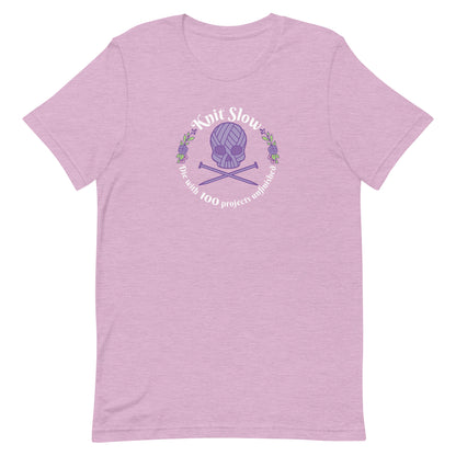 A heathered pink crewneck t-shirt featuring an image of a skull and crossbones made of yarn and knitting needles. A floral wreath surrounds the skull, along with words that read "Knit Slow, Die with 100 projects unfinished"
