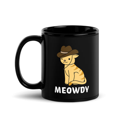 A black 11 ounce ceramic coffee mug featuring an illustration of an orange striped cat wearing a cowboy hat. The cat is winking and smiling, and text beneath him reads "Meowdy"