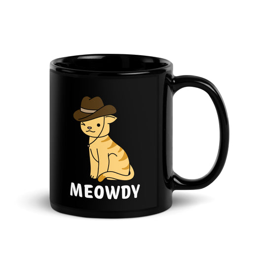 A black 11 ounce ceramic coffee mug featuring an illustration of an orange striped cat wearing a cowboy hat. The cat is winking and smiling, and text beneath him reads "Meowdy"