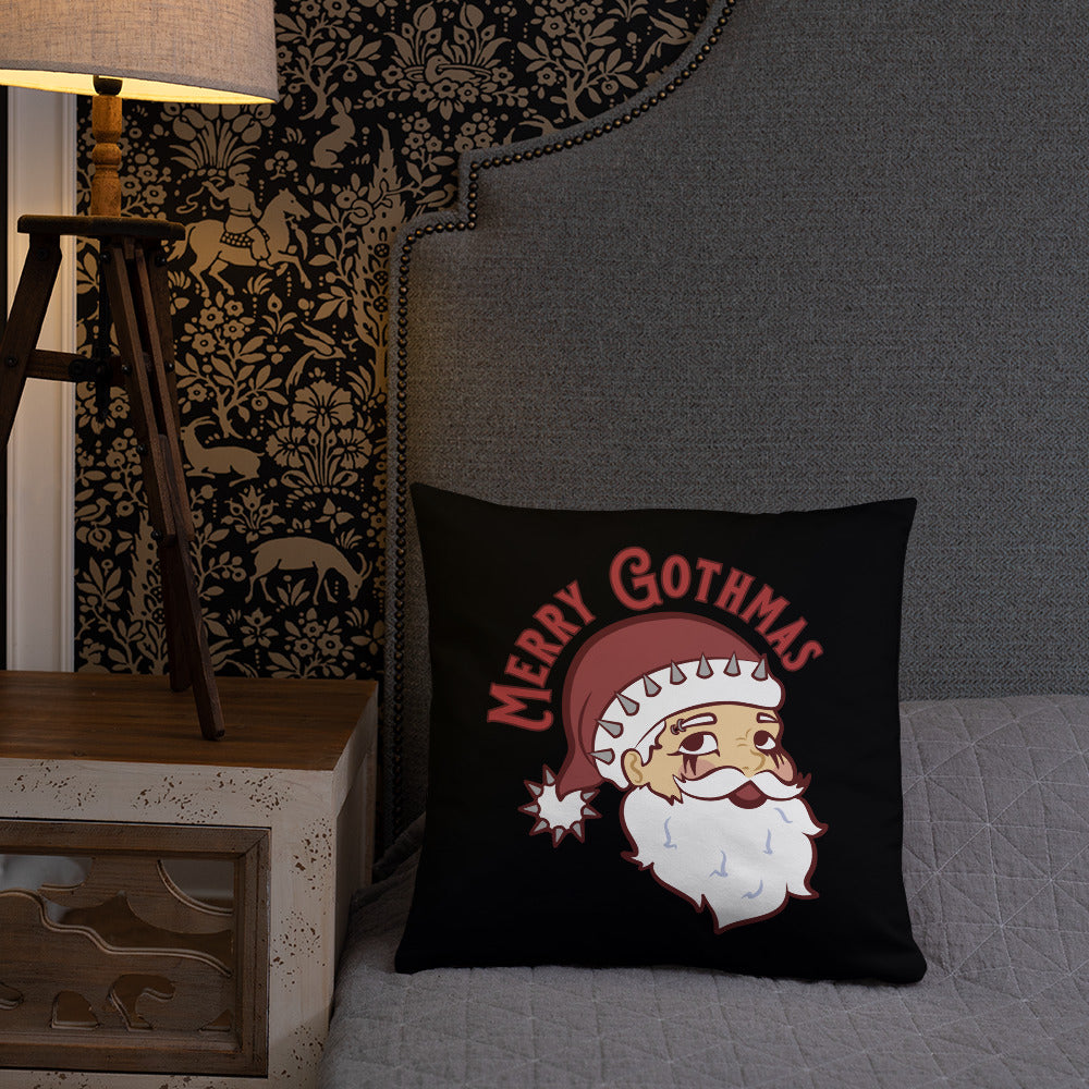A black 18" x 18" throw pillow featuring an image of Santa Claus rests on a gray bed.. Santa is wearing goth-style makeup and his hat is decorated with spikes. Text above Santa's head reads "Merry Gothmas".