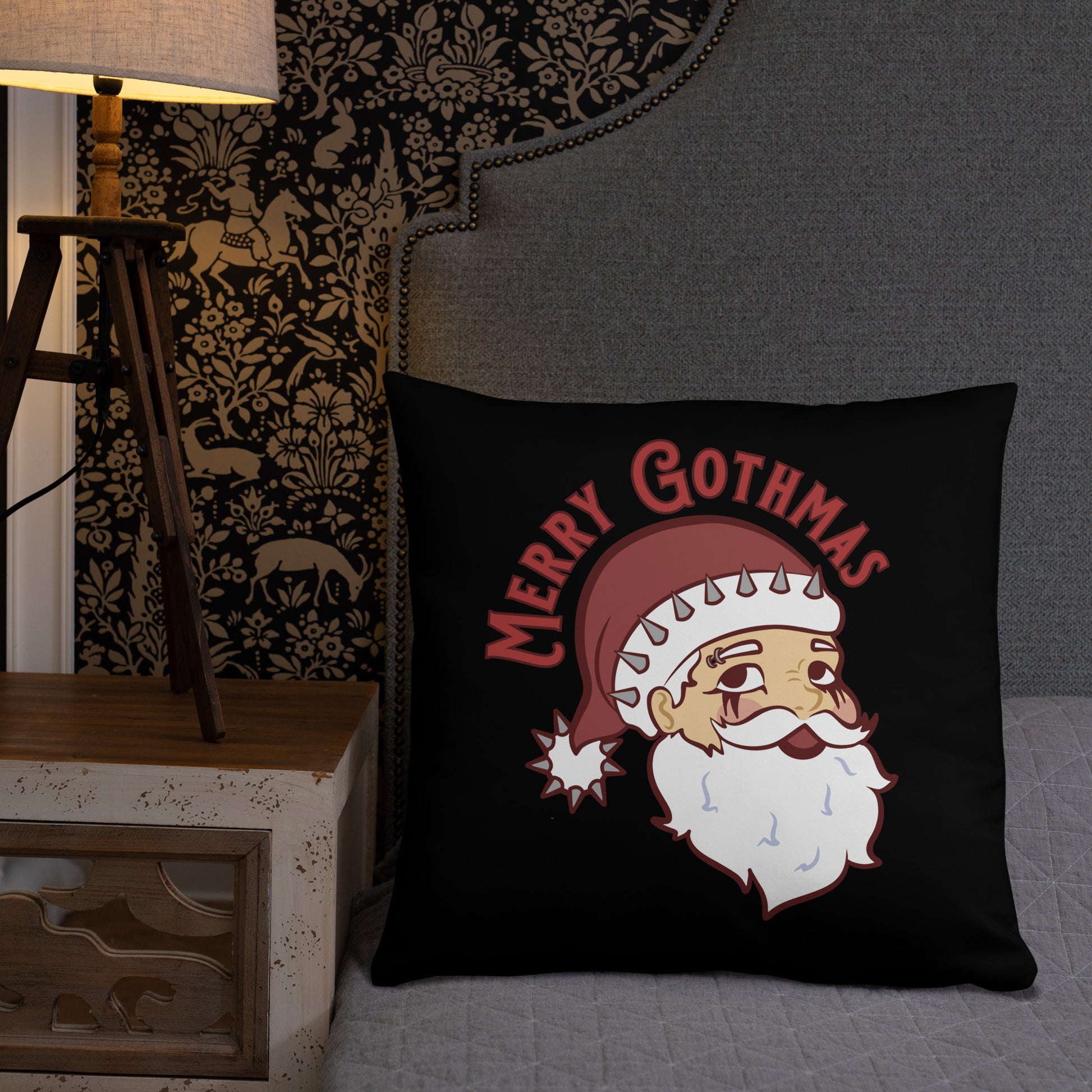 A black 22" x 22" throw pillow featuring an image of Santa Claus rests on a gray bed. Santa is wearing goth-style makeup and his hat is decorated with spikes. Text above Santa's head reads "Merry Gothmas"