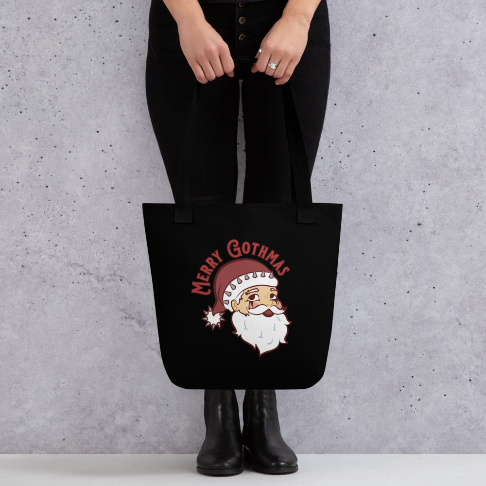 A waist-down shot of a person holding a black tote bag with black handles featuring an image of Santa Claus. Santa is wearing goth-style makeup and his hat is decorated with spikes. Text above Santa's head reads "Merry Gothmas"