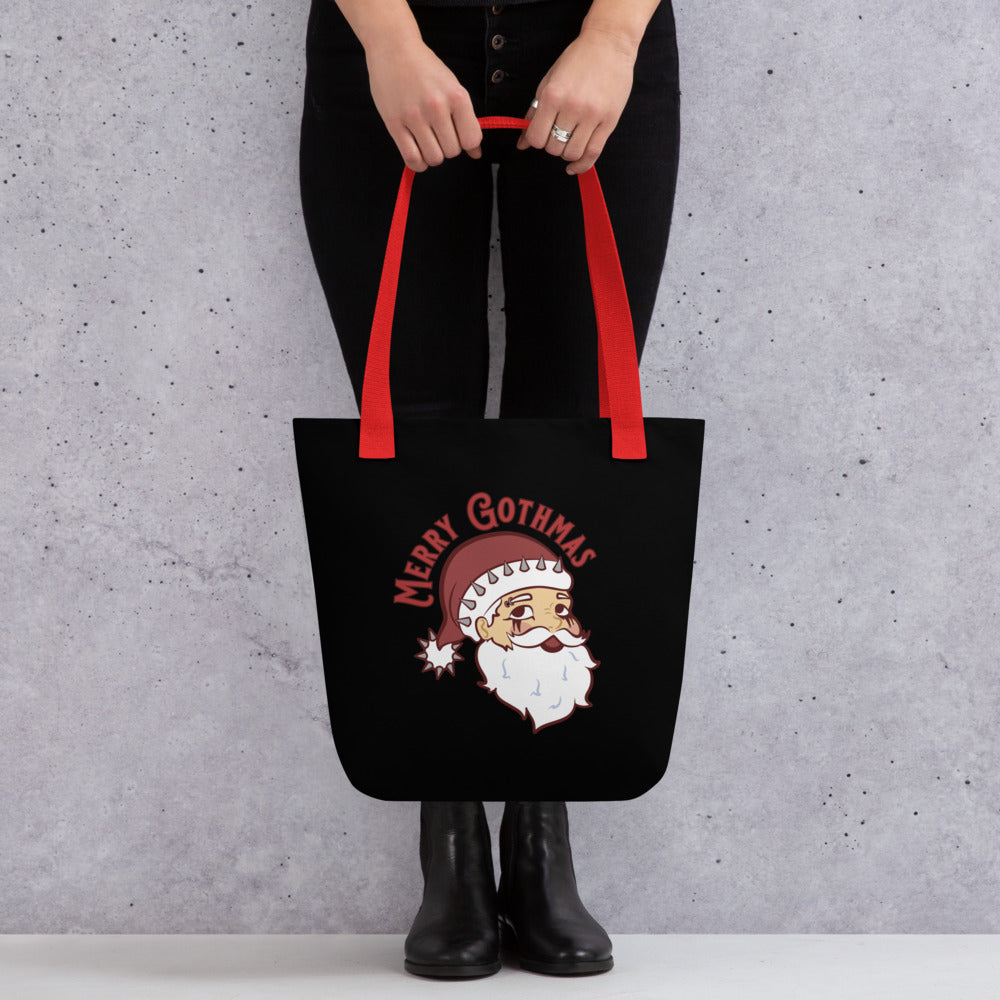 A waist-down shot of a person holding a black tote bag with red handles featuring an image of Santa Claus. Santa is wearing goth-style makeup and his hat is decorated with spikes. Text above Santa's head reads "Merry Gothmas"