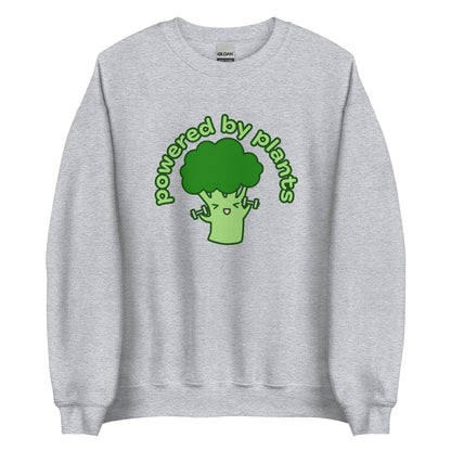 A grey crewneck sweatshirt featuring an illustration of a cartoon broccoli character. The broccoli is excitedly lifting weights, and text in an arc above the broccoli reads "powered by plants".