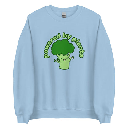 A light blue crewneck sweatshirt featuring an illustration of a cartoon broccoli character. The broccoli is excitedly lifting weights, and text in an arc above the broccoli reads "powered by plants".