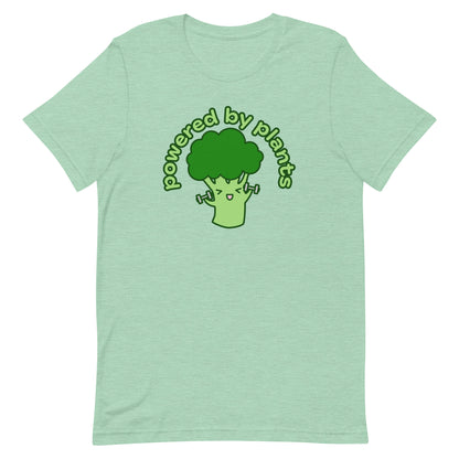 A light green crewneck t-shirt featuring an illustration of a cartoon broccoli character. The broccoli is excitedly lifting weights, and text in an arc above the broccoli reads "powered by plants".