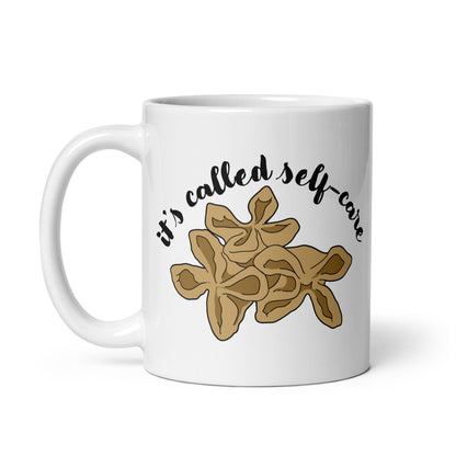 A white, 11 ounce ceramic mug featuring an illustration of three pieces of crab rangoon. Text in an arc above the crab rangoon reads "it's called self-care" in a cursive script.