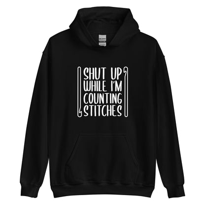 A black hooded sweatshirt featuring white text that reads "Shut up while I'm counting stitches." The text is framed by a crochet hook to the left and right.