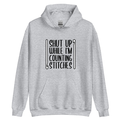 A grey hooded sweatshirt featuring black text that reads "Shut up while I'm counting stitches." The text is framed by a crochet hook to the left and right.