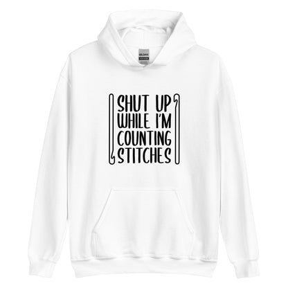 A white hooded sweatshirt featuring black text that reads "Shut up while I'm counting stitches." The text is framed by a crochet hook to the left and right.