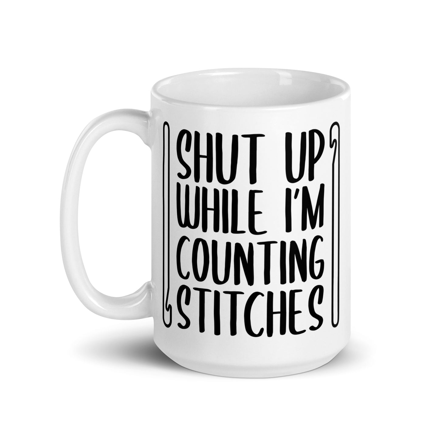 A white 15 ounce ceramic mug featuring black text that reads "Shut up while I'm counting stitches." The text is framed by a crochet hook to the left and right.