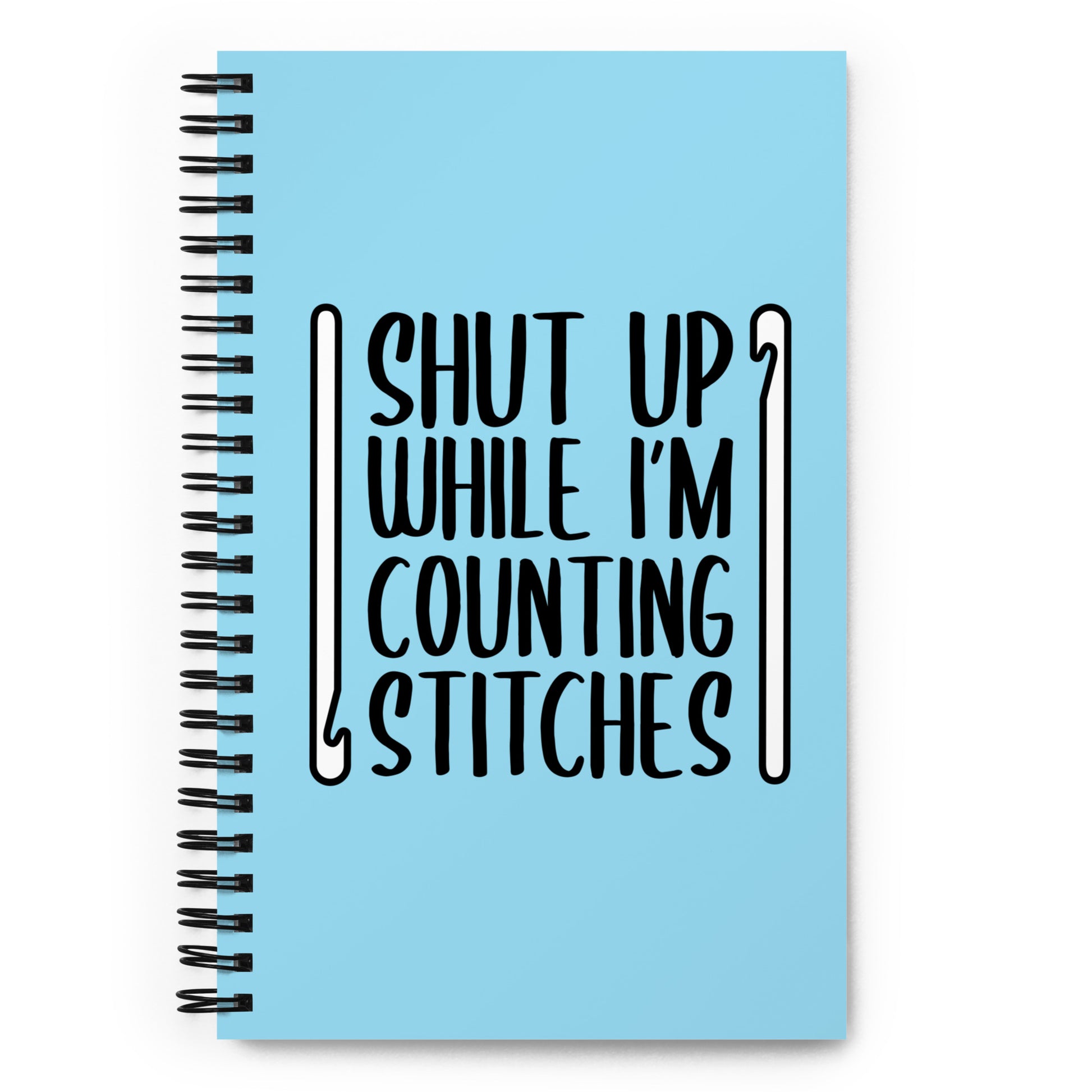 A light blue o-ring bound notebook featuring black text that reads "Shut up while I'm counting stitches." The text is framed by a crochet hook to the left and right.