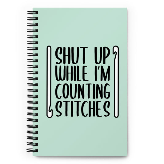 A mint green o-ring bound notebook featuring black text that reads "Shut up while I'm counting stitches." The text is framed by a crochet hook to the left and right.
