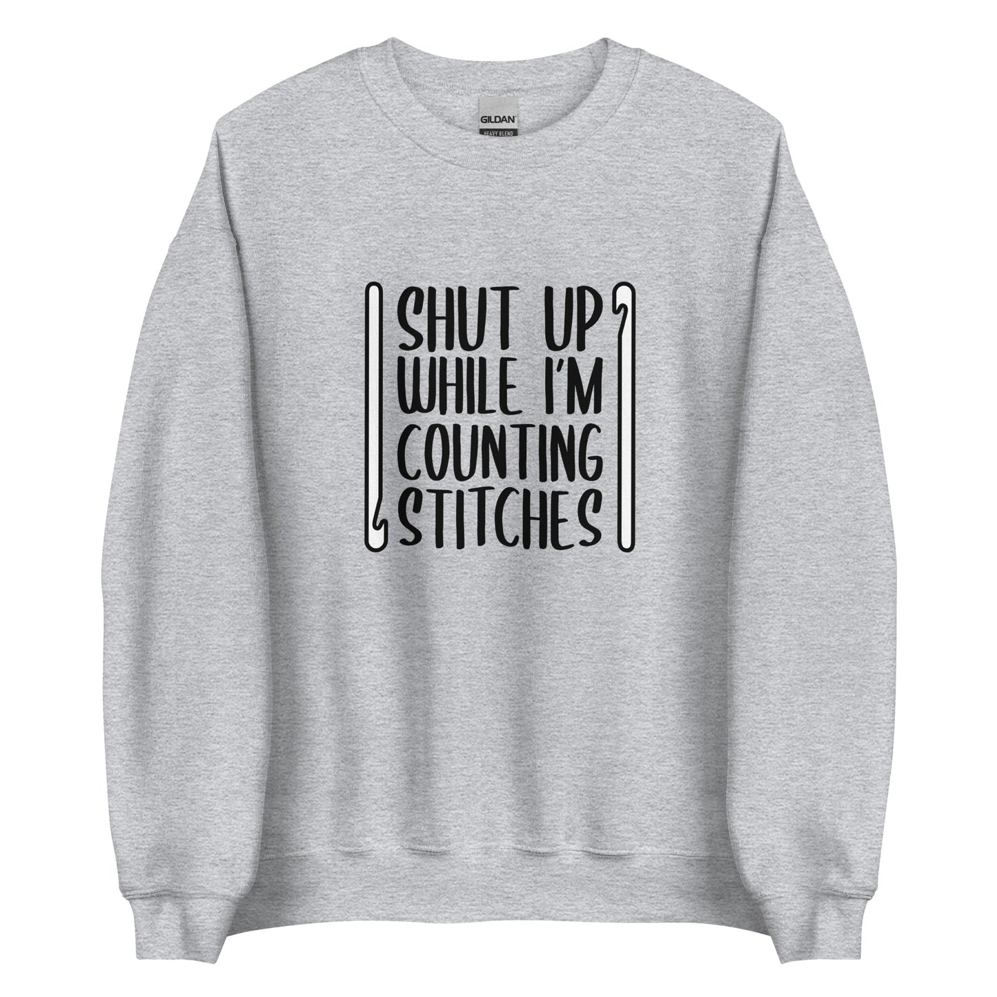 A heathered grey crewneck sweatshirt featuring black text that reads "Shut up while I'm counting stitches." The text is framed by a crochet hook to the left and right.
