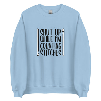 A light blue crewneck sweatshirt featuring black text that reads "Shut up while I'm counting stitches." The text is framed by a crochet hook to the left and right.