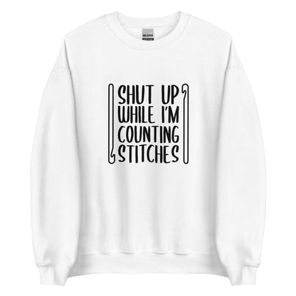A white crewneck sweatshirt featuring black text that reads "Shut up while I'm counting stitches." The text is framed by a crochet hook to the left and right.