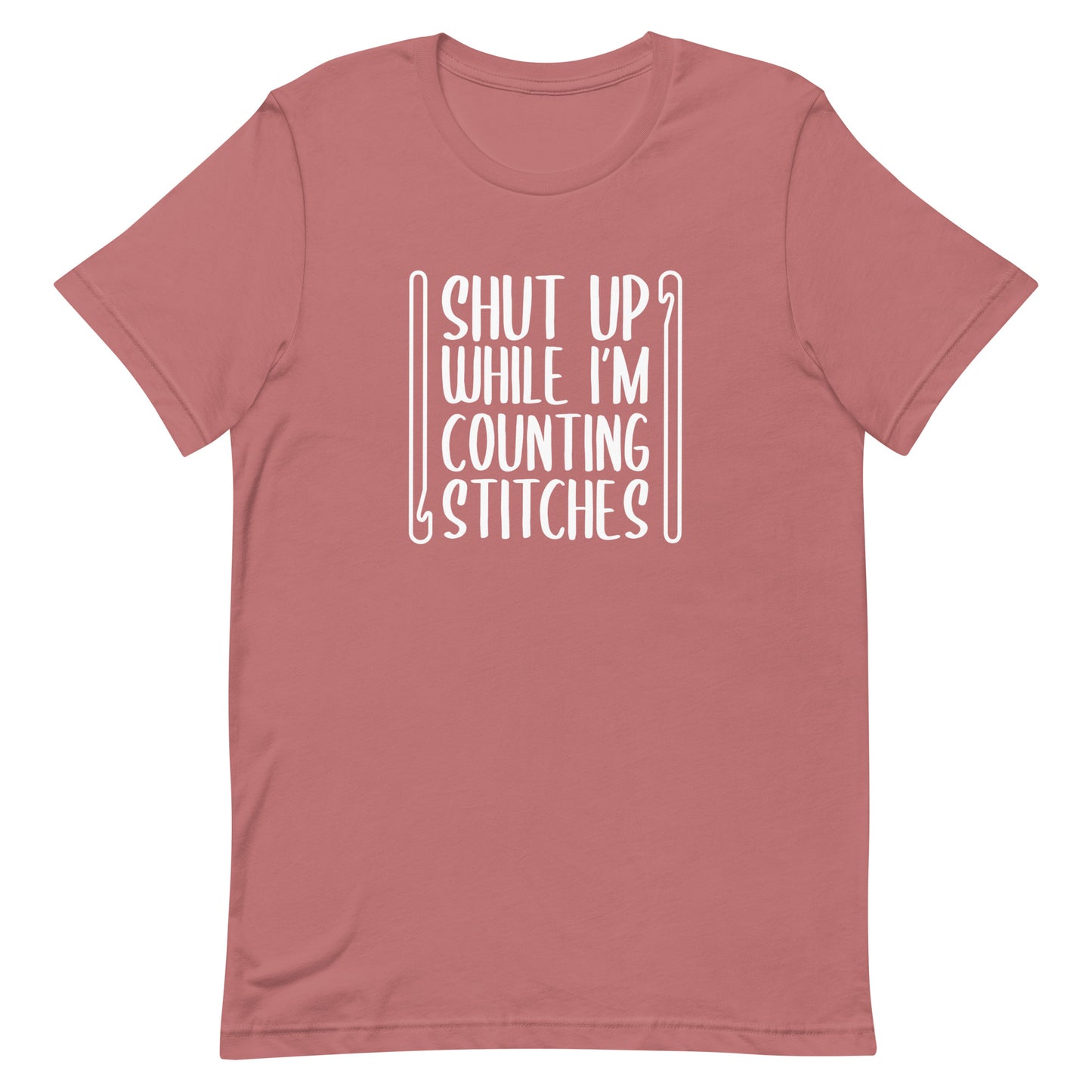 A dusky pink crewneck t-shirt featuring white text that reads "Shut up while I'm counting stitches." The text is framed by a crochet hook to the left and right.