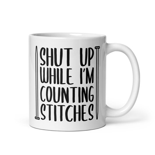 A white 11 ounce ceramic mug with black text surrounded by two knitting needles. The text reads "Shut up while I'm counting stiches".
