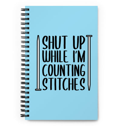A blue wirebound notebook with black text surrounded by two knitting needles. The text reads "Shut up while I'm counting stiches".