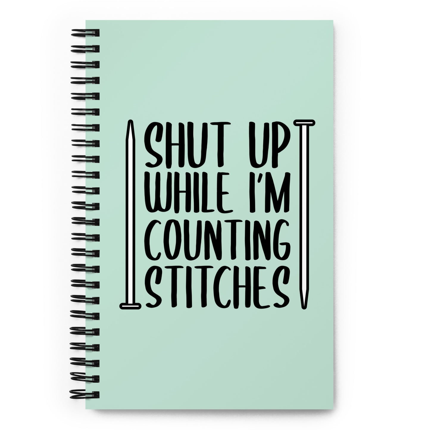 A light mint green wirebound notebook with black text surrounded by two knitting needles. The text reads "Shut up while I'm counting stiches".