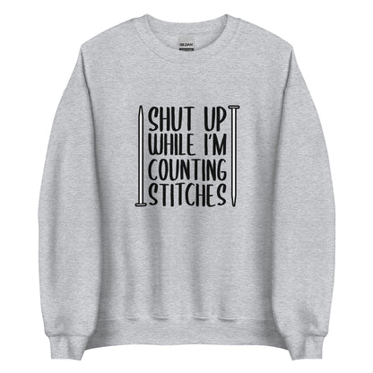 A grey crewneck sweatshirt with black text surrounded by two knitting needles. The text reads "Shut up while I'm counting stiches".