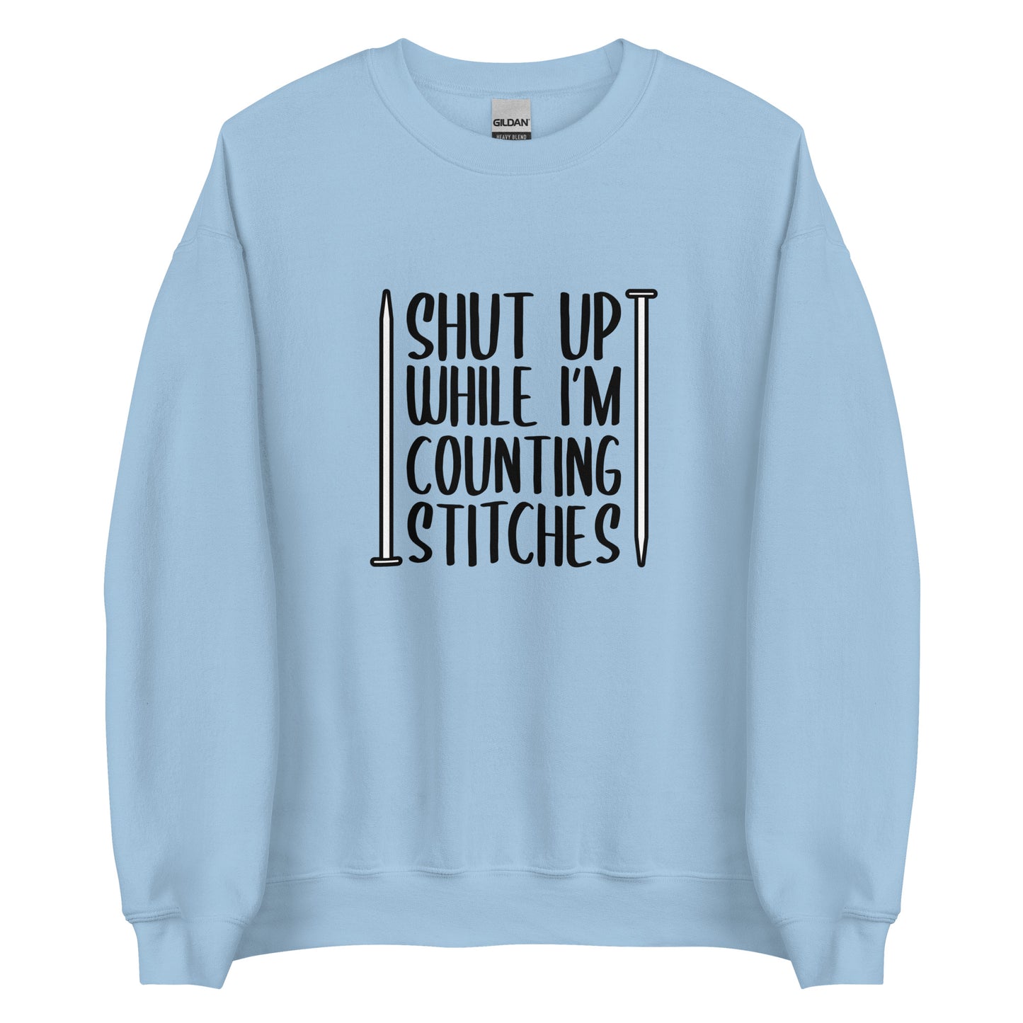 A light blue crewneck sweatshirt with black text surrounded by two knitting needles. The text reads "Shut up while I'm counting stiches".
