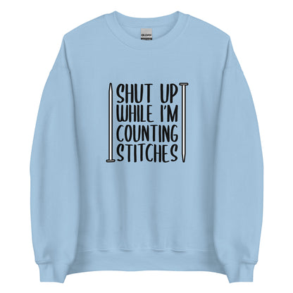 A light blue crewneck sweatshirt with black text surrounded by two knitting needles. The text reads "Shut up while I'm counting stiches".