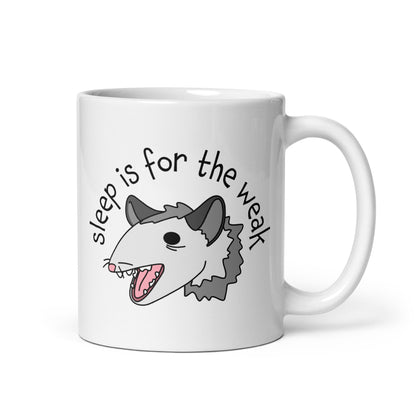 A white 11 ounce ceramic coffee mug featuring an illustration of an opossum with its mouth open, as if it was yelling. Text alongside the opossum reads "sleep is for the weak"