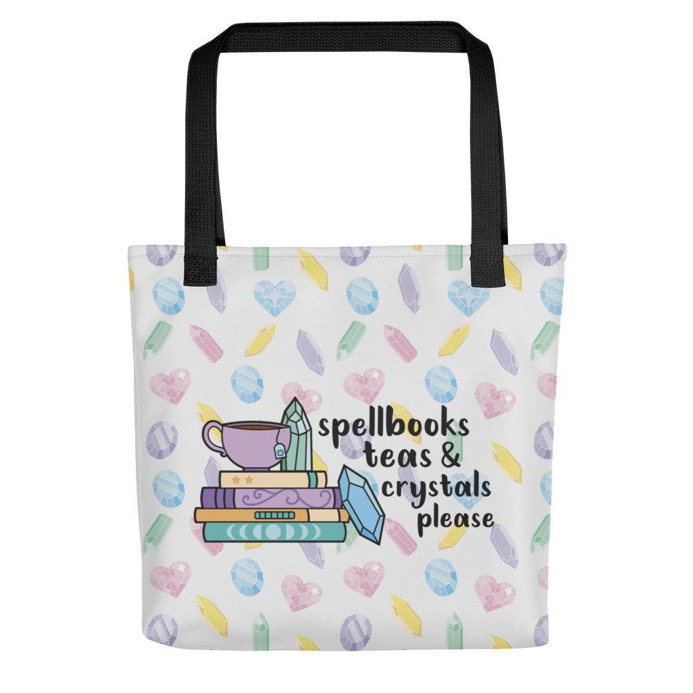 A white totebag with black handles, decorated with a pattern of pastel-colored crystals. An illustration on the bag shows a stack of spellbooks with a teacup on top and crystals scattered around the books. Text next to the image reads "Spellbooks, teas & crystals please"